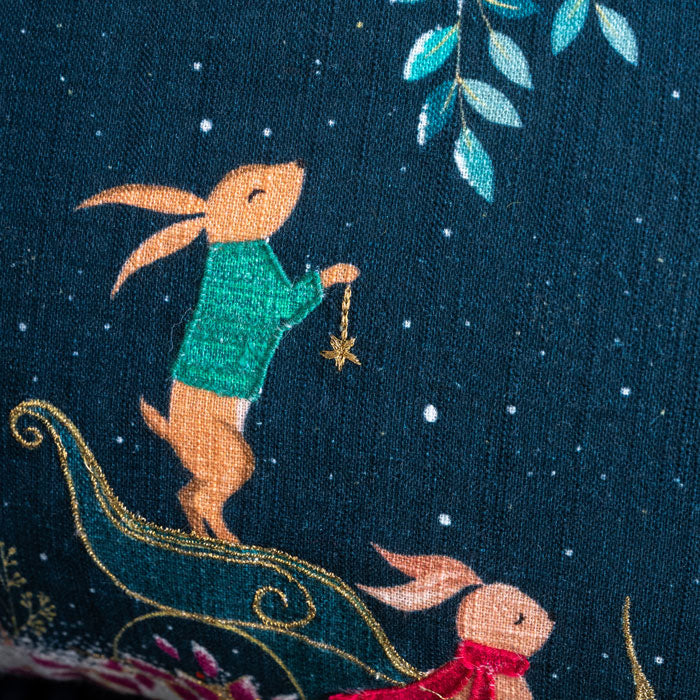 Embroidered scene of a rabbit in a green sweater holding a star, with another rabbit in a red scarf in a sleigh on a dark blue cushion.