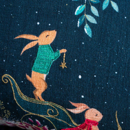 Embroidered scene of a rabbit in a green sweater holding a star, with another rabbit in a red scarf in a sleigh on a dark blue cushion.