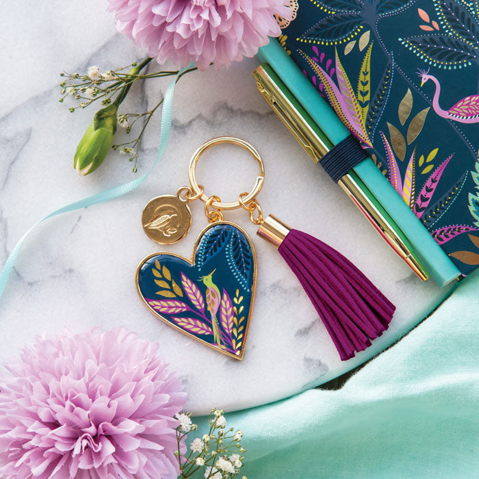 Heart-shaped keychain with tassel on a marble surface with flowers and a notebook.