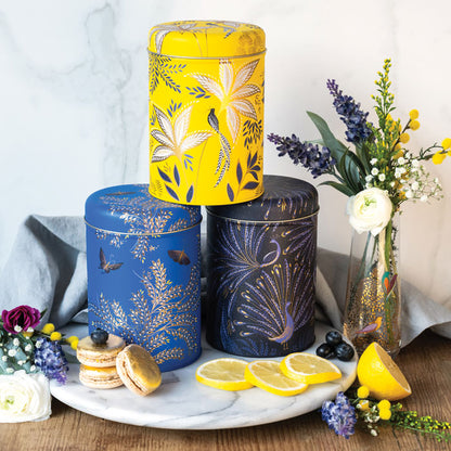 Three round tin storage caddies tacked, in different colours yellow, cobalt and indigo, each with different floral and bird designs.