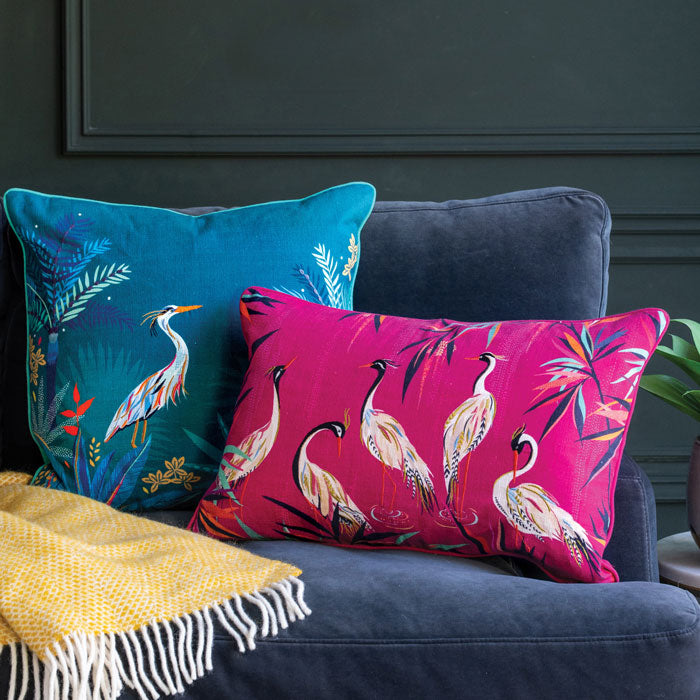 Two decorative pillows, one teal heron and one pink heron on a blue sofa against a dark wall.