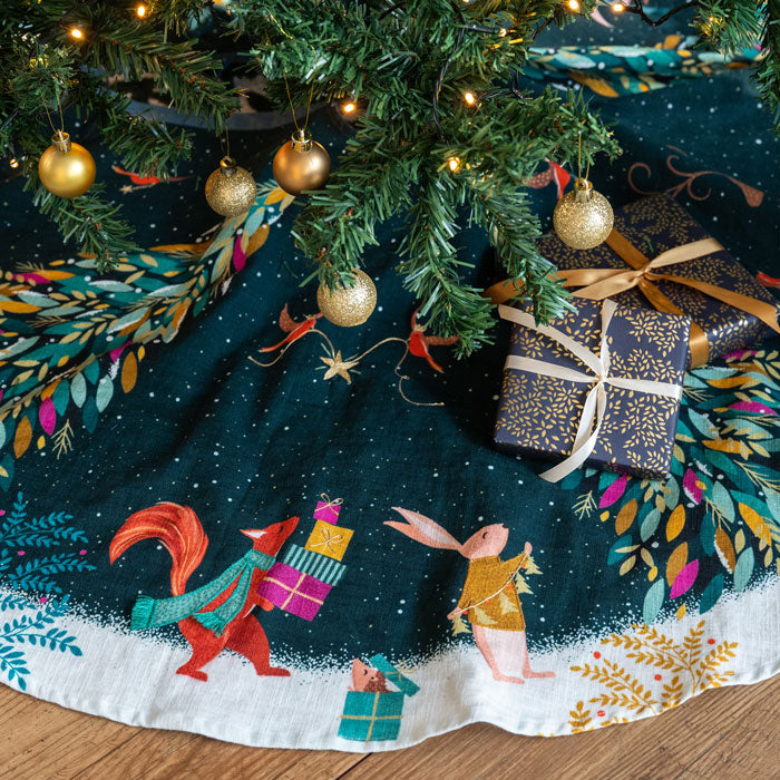 Decorative Christmas tree skirt with animal illustrations and a gift, under a decorated Christmas tree.