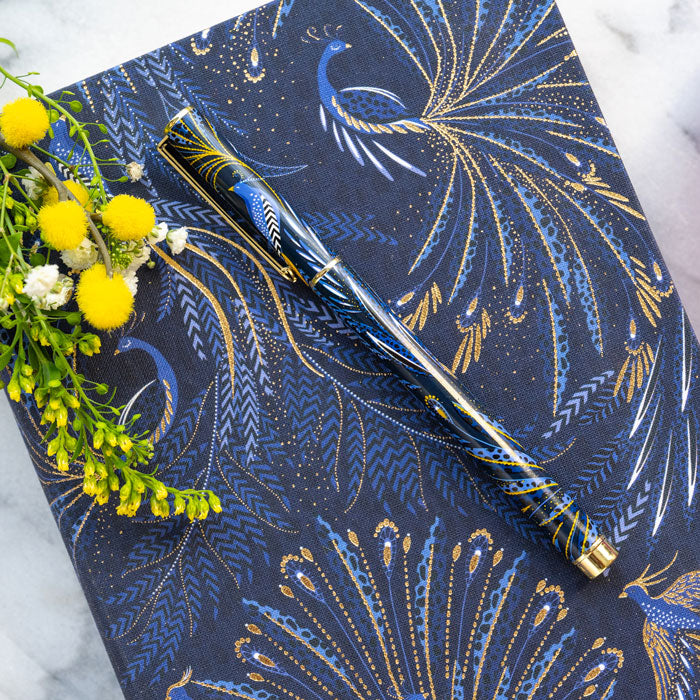 Ballpoint pen presented in box, with peacock feathers design in indigo and gold set on matching notebook.