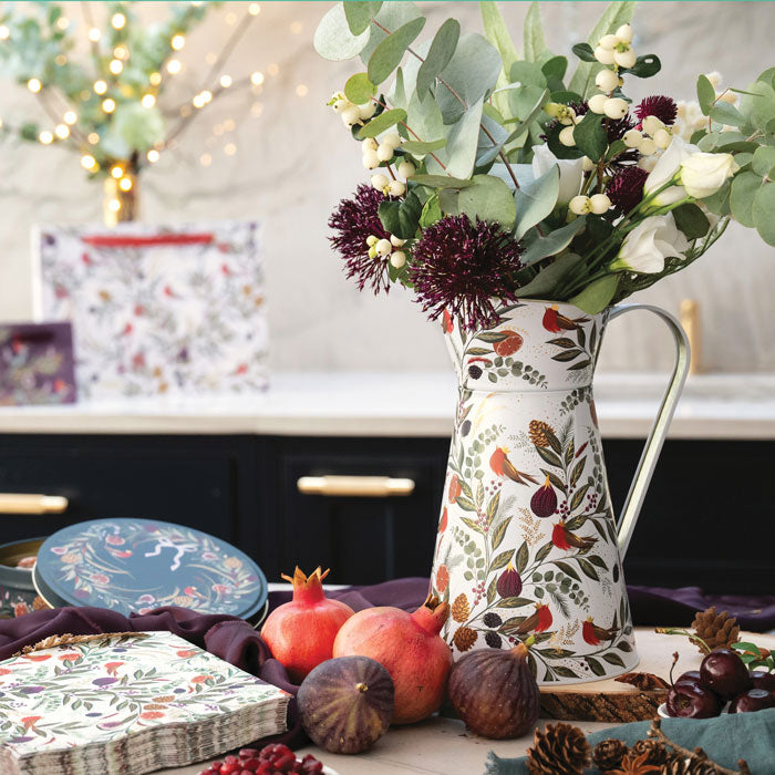 A white tin flower jug with a floral festive  design of robins, figs, red berries, green leaves, filled with eucalyptus and white flowers, set on a table with pomegranates, figs, and gift boxes in a kitchen setting.