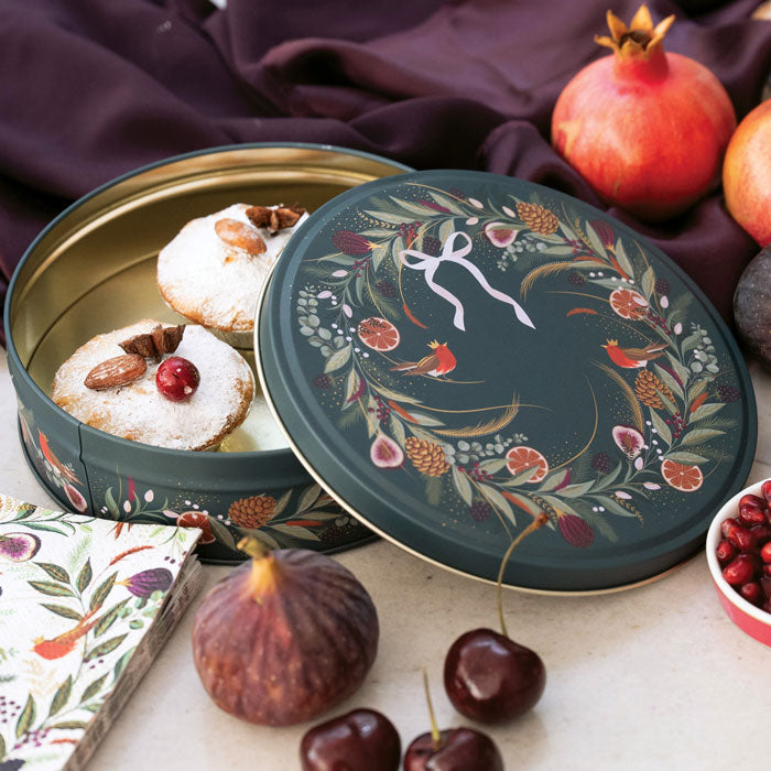A round decorative metal tin, with the lid leaning beside it. The tin has a dark green floral festive wreath design with robins, figs, and citrus motifs.