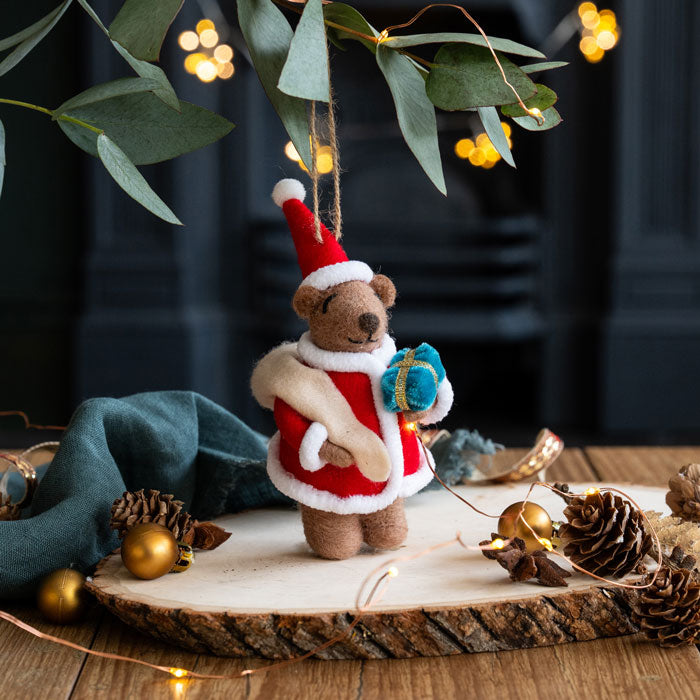 Small teddy bear in a Santa outfit holding a gift, surrounded by Christmas decorations on a wooden surface.
