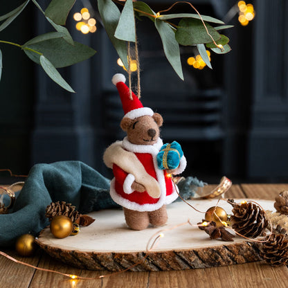 Small teddy bear in a Santa outfit holding a gift, surrounded by Christmas decorations on a wooden surface.