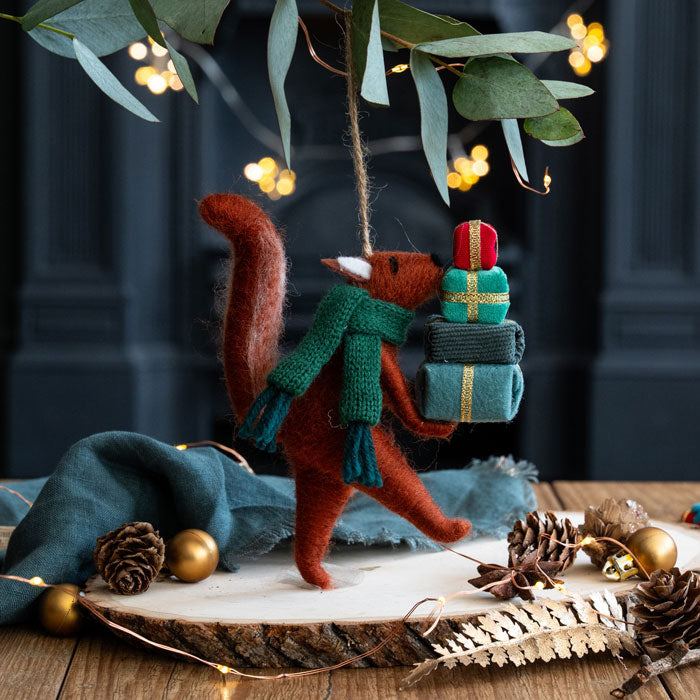 Felted fox ornament with a green scarf and gift, hanging from eucalyptus leaves on a wooden surface with decorative items.