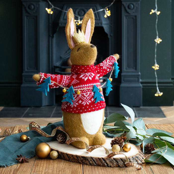 Felt rabbit figuring wearing a red sweater with Christmas tree lanyard decorations in front of a fireplace.