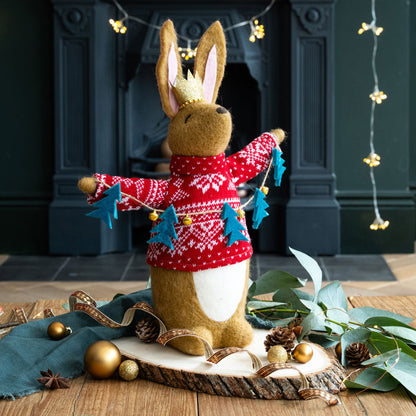 Felt rabbit figuring wearing a red sweater with Christmas tree lanyard decorations in front of a fireplace.