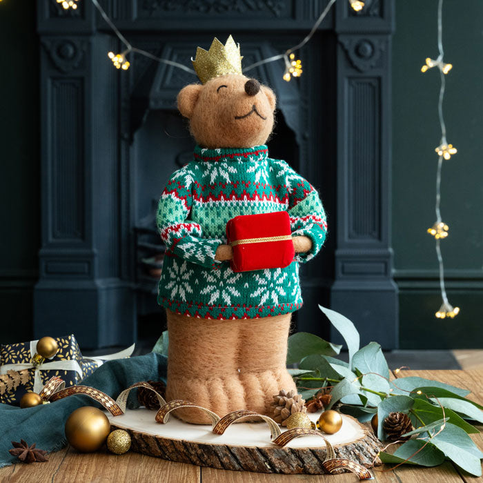 Teddy bear wearing a festive sweater and holding a gift in front of a fireplace with decorative lights.