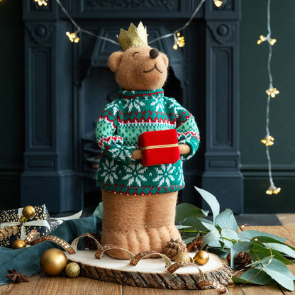 Teddy bear wearing a festive sweater and holding a gift in front of a fireplace with decorative lights.