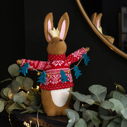 Felt rabbit figurine wearing a red sweater with Christmas trees lanyard, standing on a dark background with greenery.