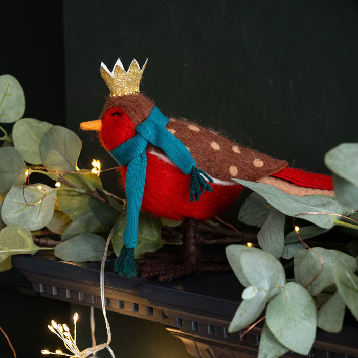 Decorative robin with a crown and blue scarf on a dark background with greenery and lights.