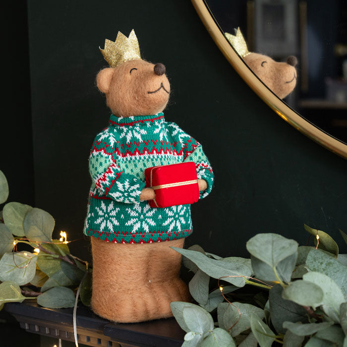 Teddy bear wearing a festive sweater and holding a gift, standing in front of a mirror with greenery around.